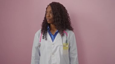Doktor, üniformalı Afro-Amerikan bir kadın ciddi bir yüz ve doğal bir ifadeyle izole edilmiş pembe arka planda kameraya bakıyor.