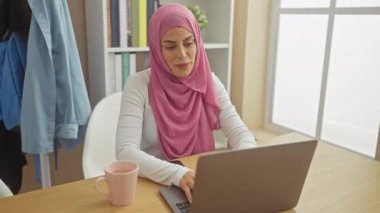 Pembe tesettürlü bir kadın evinde dizüstü bilgisayarı, akıllı telefonu ve masanın üstünde bir fincanla esniyor..