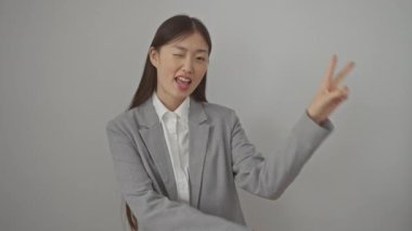 Ceket giyen neşeli genç Çinli kadın, izole edilmiş beyaz bir arka planda duruyor, muzaffer bir 'V' sembolünü iki parmakla gösteriyor, gülümsüyor ve kameraya göz kırpıyor.