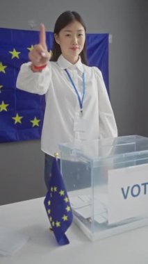 Rozeti olan Asyalı bir kadın. Evinde bir bayrak ve oy sandığının önünde baş parmak kaldırıyor..