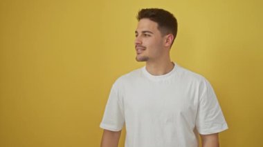 Beyaz tişörtlü neşeli, genç İspanyol adam, iki eliyle kenarı göstererek, sarı izole bir arka plana karşı duruyor, sizi parlak bir gülümsemeyle karşılıyor.!