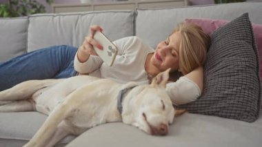 Genç, beyaz bir kadın, uyuyan köpeğiyle evde bir koltukta rahatlar. Akıllı telefondaki videoyu izlerken neşeyle gülümser..