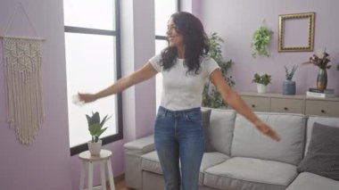 Neşeli bir kadın, modern bir oturma odasında, bitkilerle, makrome bir duvarla ve şık mobilyalarla duruyor..