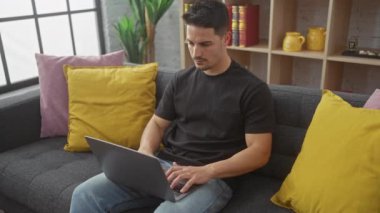 Yakışıklı İspanyol adam dizüstü bilgisayarı kapatıyor ve rahat bir oturma odasında kameraya gülümsüyor.