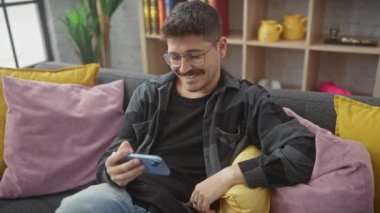 Gülümseyen İspanyol bir adam, gözlük takıyor ve rahat bir şekilde akıllı telefon kullanıyor renkli yastıklarla rahat bir oturma odasında dinlenirken..