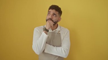 Önlük giymiş düşünceli genç İspanyol bir adam, elleri çenesinde, izole edilmiş sarı arka planda, ciddi bir bakışla ciddi bir soru ve fikir üzerinde kafa yoruyor.