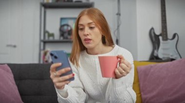 Elinde pembe kupa tutan, şaşırmış, genç bir kadın rahat bir oturma odasında akıllı telefonuna tepki veriyor..