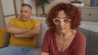 Orta yaşlı bir çift oturma odasında oturuyor, üzgün görünüyorlar ve göz temasından kaçınıyorlar, ilişkilerindeki gerilimi ve hayal kırıklığını vurguluyorlar..