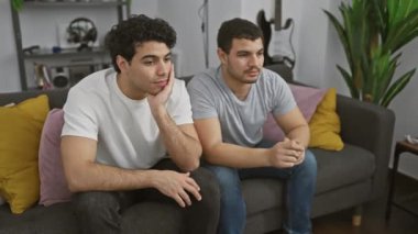 Rahat bir oturma odasında gri bir kanepede oturan iki genç adam, yansıtıcıdan neşeye çeşitli duygular sergiliyorlar.