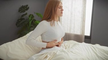 Kederli, beyaz, kızıl saçlı bir kadın yatak odası ortamında bir yatağa oturmuş karnını sıkıyor..