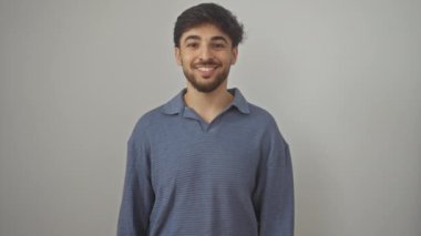 Mutlu, genç Arap adam kendine güvenerek duruyor, dişlek bir gülümseme ile pozitiflik yayıyor, izole beyaz bir arka planda neşeli bakıyor.