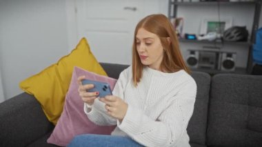 Genç bir kadın evde vakit geçirmekten zevk alıyor, rahat bir oturma odasında akıllı telefonuyla geziniyor..