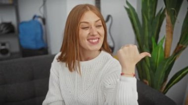 Evdeki neşeli, genç, kızıl saçlı kadın, başparmağıyla sevinçten havalara uçuyor, süveter giyiyor ve ağzı açık gülümsüyor.