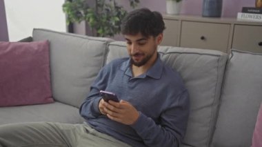 Genç Arap adam gururlu bir zaferi kutluyor, heyecanlı ifadeler, kollarını kaldırıyor, akıllı telefondaki başarılar evde koltukta otururken.
