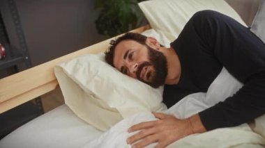 Sakallı bir adam uzanıyor, gülüyor ve yatak odasında yastıkla oynuyor..