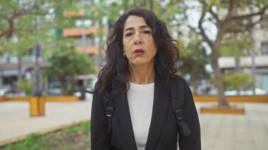 Orta yaşlı, kıvırcık saçlı, İspanyol bir kadın, hareketli bir şehir parkında spor ceket giyerek düşünceli bir şekilde dikiliyor..