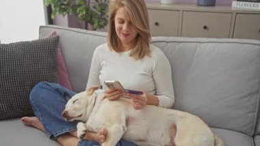 Elinde akıllı telefon ve kredi kartıyla uyuyan köpeğiyle koltukta dinlenen gülümseyen bir kadın.