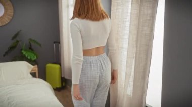 Gündelik kıyafetli genç bir kadın modern bir yatak odasında pencerenin kenarındaki bir yatağa oturmuş, dalgın dalgın dalgın bakıyor..