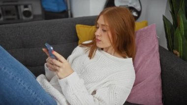 Rahat bir kadın evdeki kanepede uzanıyor, akıllı telefonunu sıradan bir oturma odasında kullanıyor..