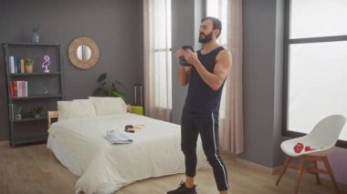 Orta yaşlı sakallı bir adam modern bir yatak odasında kettlebell çömelmesi yapıyor, evde spor yapmaya odaklanıyor.