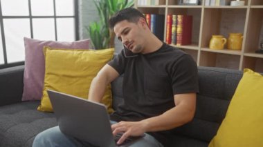 İspanyol adam evinde rahat bir oturma odasındaki kanepede dizüstü bilgisayar ve akıllı telefonla birden fazla iş yapıyor..