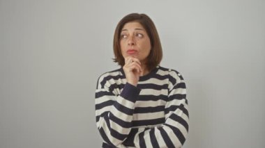 Düşünceli orta yaşlı İspanyol kadın çizgili kazak giyiyor, ciddi ciddi kafa karıştırıcı sorular soruyor, çenesi kapalı beyaz arka plan