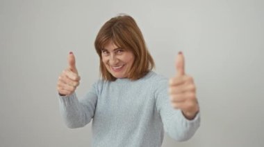 Süveter giyen neşeli orta yaşlı kadın, izole edilmiş beyaz arka planda ışıldayan bir gülümseme ile muzaffer, baş parmak işareti yapıyor. Başarıyı kutluyor, pozitiflik ve mutluluk yayıyor..