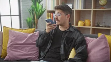 Kafası karışmış, gözlüklü ve bıyıklı bir İspanyol adam renkli yastıklı, rahat bir oturma odasında akıllı telefonunu kullanıyor..