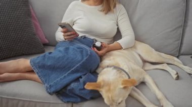 Genç yetişkin bir kadın, elinde akıllı telefon ve şişeyle evcil köpeğiyle kanepede dinlenir..
