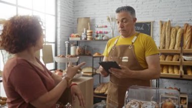 Kadın, fırında tablet kullanan bir adamla konuşuyor. Raflar ekmek ve hamur işleriyle dolu. Modern bir ev ortamında.