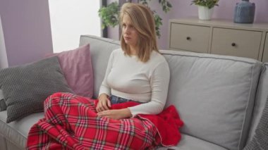 Rahat bir oturma odasında kırmızı bir battaniyeyle kanepede oturan endişeli genç kadın..