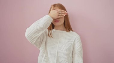 Şok edici bir bakış! Genç, kızıl saçlı, utangaç ve utangaç bir kadın, elleriyle yüzünü gizliyor, izole edilmiş pembe arka planda parmaklarının arasından dikizliyor.