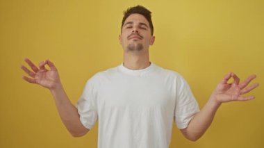 Gözleri kapalı duran, beyaz tişörtlü, sakin, genç İspanyol adam etkileyici bir yoga meditasyon hareketi yapıyor. Gevşemeye daldırılmış, canlı sarı bir arkaplanda izole edilmiş..