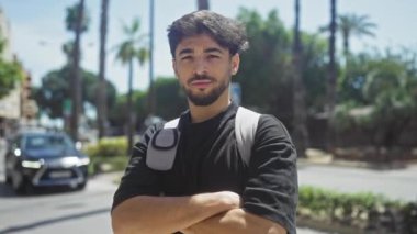 Şehir caddesinde duran sakallı, kendine güveni tam genç bir adam, kolları bağlı, gündelik tarzda, gündüz vakti..