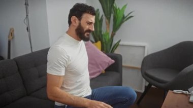 Beyaz tişörtlü yakışıklı, nefret dolu sırt ağrısı çeken, güçlü bir elle sırtına dokunan, rahat oturma odasında kas zedelenmesi belirtileri gösteren orta yaşlı bir adam..