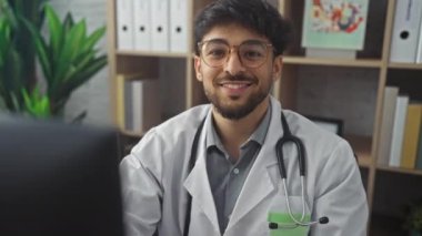 Gözlüklü neşeli bir genç klinikte steteskoplu laboratuvar önlüğü giyiyor..