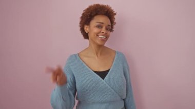 Afro-Amerikalı kadın kazak giyip gülümsüyor ve kameraya bakıp zafer işareti yapıyor. İki numara. izole edilmiş pembe arkaplan