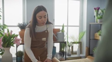 Kahverengi önlüklü, gülümseyen bir kadın, arka planda çeşitli bitkiler bulunan kapalı bir çiçekçide müşteriye çiçek verir..