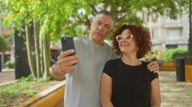 Şehir parkında selfie çeken orta yaşlı bir çift güneşli bir günde birlikte gülüyor ve dışarıda eğleniyorlar. Gri gömlekli bir adam ve kıvırcık saçlı gözlüklü bir kadın.