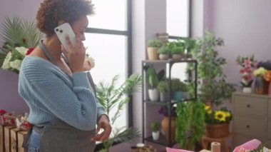 Kapalı bir çiçekçide renkli çiçeklerle telefonda sohbet eden Afrikalı Amerikalı bir kadın.