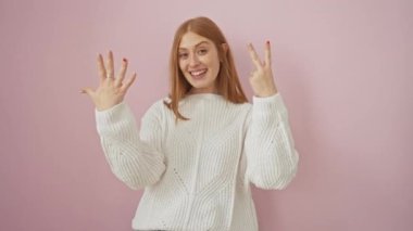 Pembe izole edilmiş arka planda güvenle duran parlak kızıl saçlı kadın parmaklarını yukarı kaldırarak yedi numaranın sembolünü mutlulukla gösteriyor.
