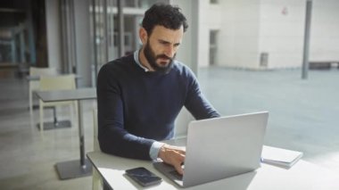 Orta yaşlı sakallı bir adam, modern bir ofis ortamında dizüstü bilgisayarla çalışırken rahatsız olur..