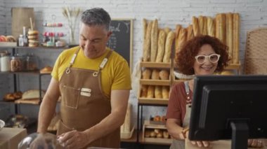 Kadın ve erkek bir fırında birlikte çalışıyorlar. Adam tepsi örtüsünü tutuyor ve kadın bilgisayar kullanıyor. Etrafı çeşitli ekmek ve hamur işleriyle çevrili.