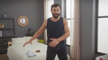 Orta yaşlı, sakallı bir adam gündelik giysiler içinde modern bir yatak odasında poz veriyor..