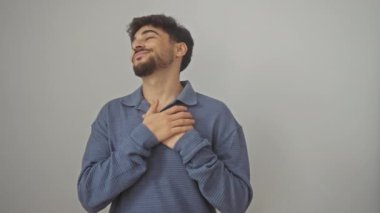 Genç Arap adam, elleri göğsünde, sağlık jestlerini tasvir ediyor, izole edilmiş beyaz bir arkaplanda gülümsüyor, minnettar bir yüzü ve kapalı gözleri var..