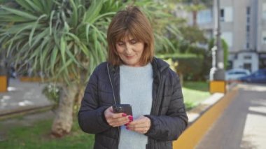 Açık havada akıllı telefon kullanan, kısa saçlı ve manikürlü manikürlü bir ceketle gülümseyen olgun bir kadın..