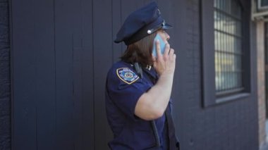 Üniformalı olgun kadın polis memuru şehir caddesinde akıllı telefonla iletişim kuruyor..