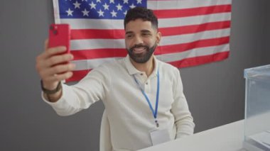 Sakallı yakışıklı İspanyol bir adam arka planda bayrağı olan bir Amerikan seçim odasında selfie çekiyor.