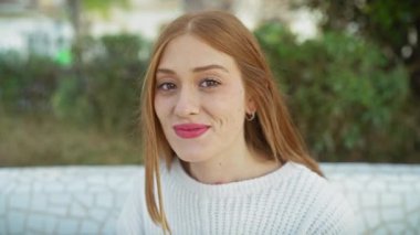 Beyaz kazaklı, gülen kızıl saçlı kadın açık havada sakin bir anın tadını çıkarıyor..