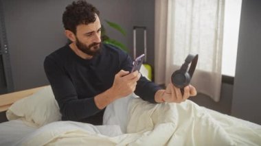 Düşünceli bir adam yatak odasında kulaklıkla akıllı telefonu inceliyor. Merakını ya da şaşkınlığını dile getiriyor..